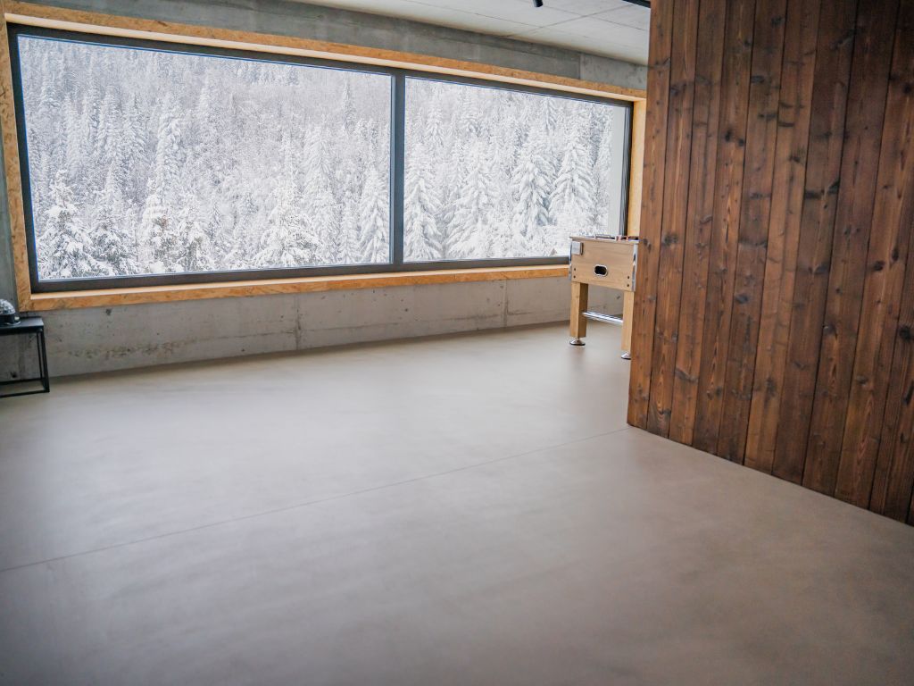 Mikrocement wady i zalety: Podłoga z szarego mikrocementu w pokoju rozrywkowym z dużymi oknami z widokiem na las.